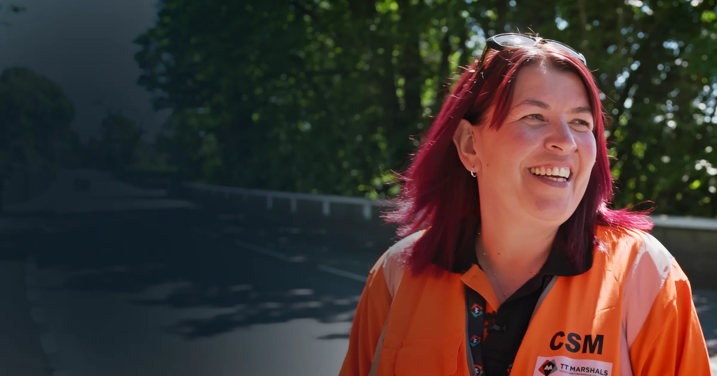 Smiling person in an orange jacket stands outdoors, trees in background.